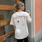 Person wearing a white t-shirt with 'Cats & Wine' design, holding a green cup, standing outside a building.